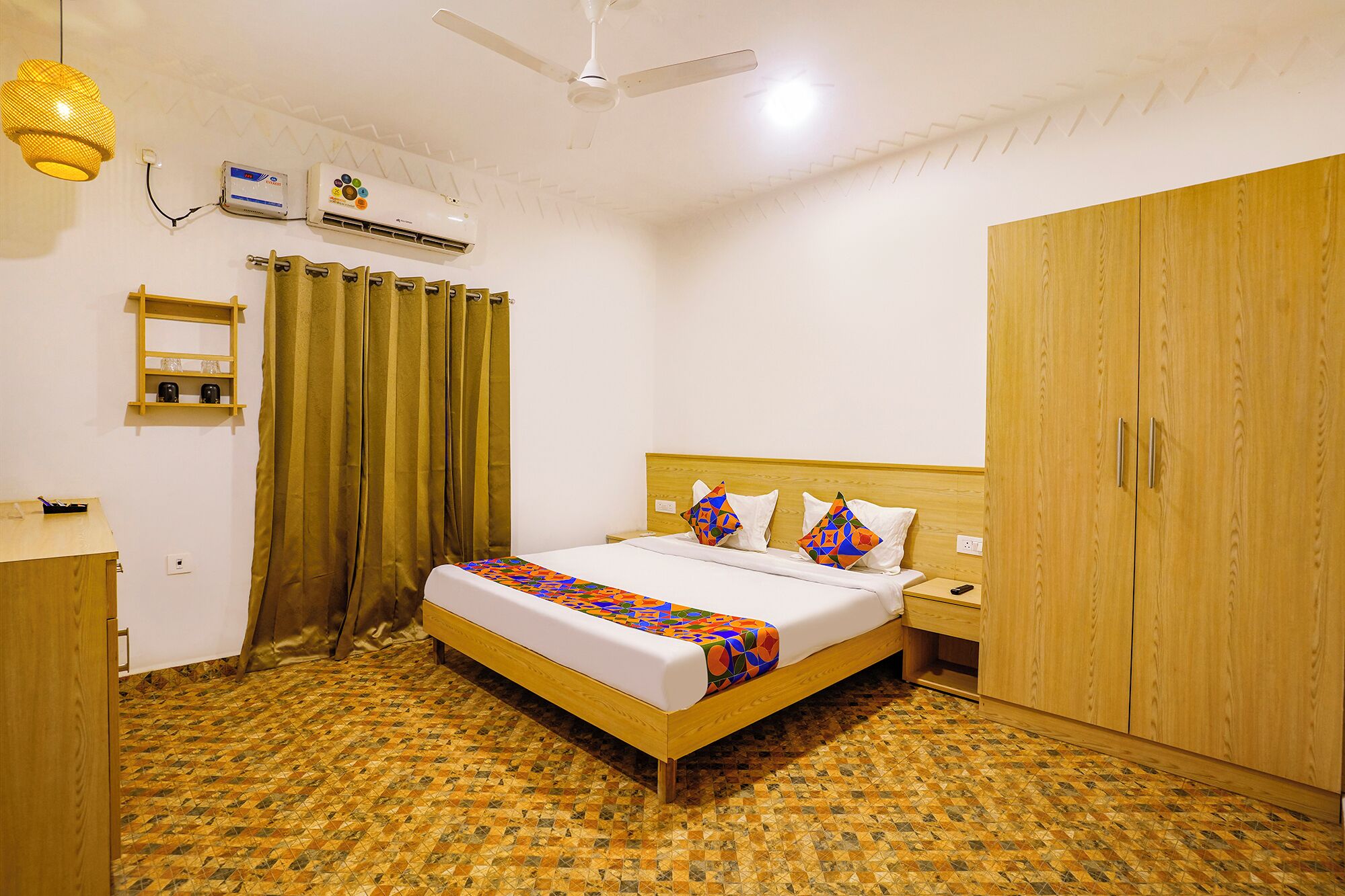 A hotel room with a bed, wooden wardrobe, a desk, a chair, a nightstand, an air conditioning unit, and a ceiling fan.