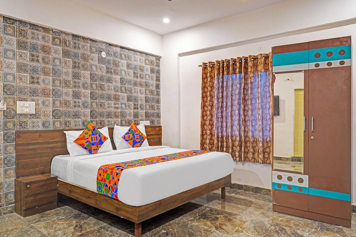 Un dormitorio con pared estampada, una cama con almohadones coloridos, una mesita de noche de madera, un armario con espejo y una cortina con diseño circular.