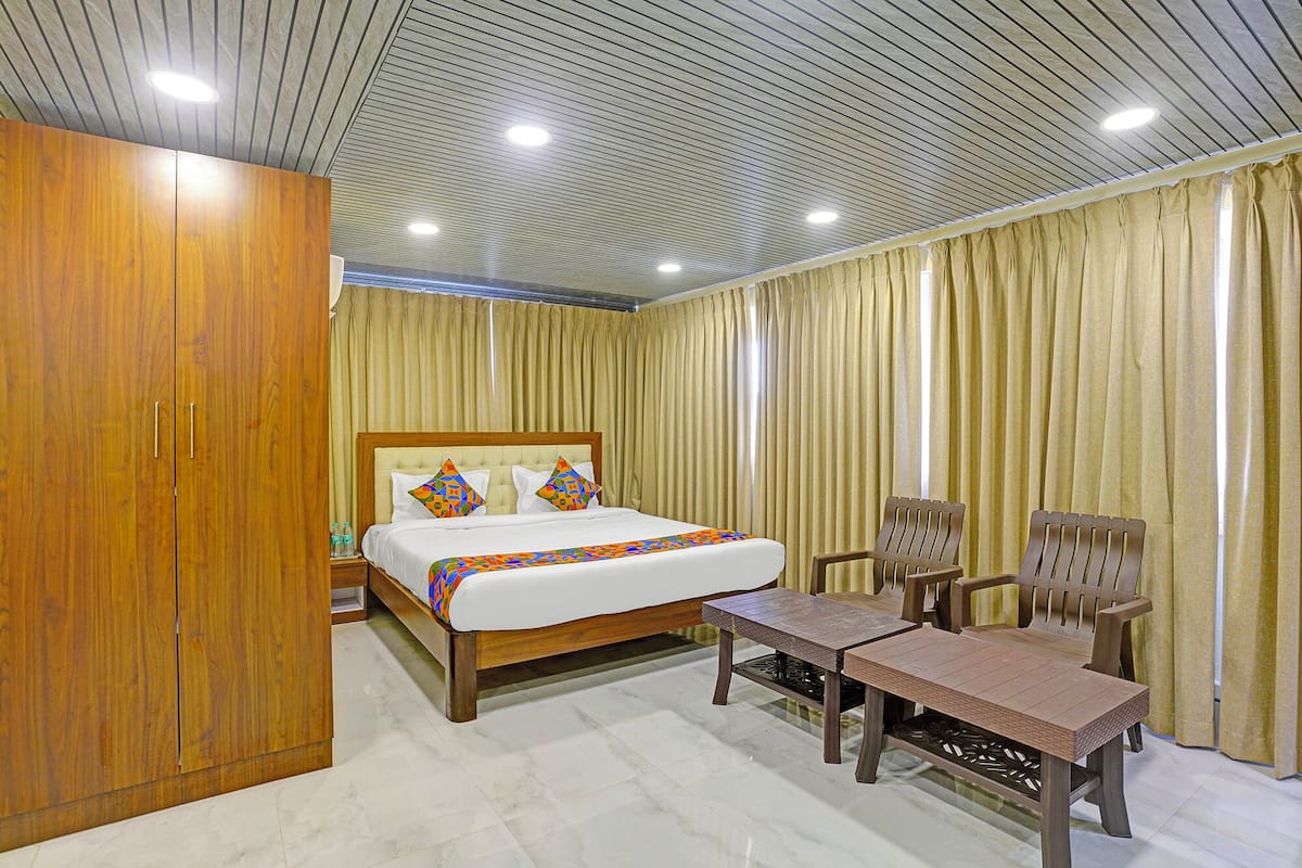 A hotel room with a wooden wardrobe, a bed with colorful pillows, a wooden bench, and a large window with curtains.