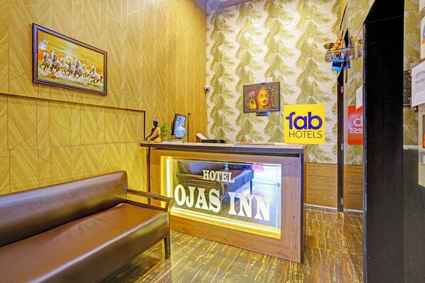 A reception desk with a sign reading 'HOTEL' and 'JAS INN' in a hotel lobby with patterned wallpaper and a brown leather bench.