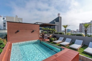 Ein Pool auf dem Dach mit Liegestühlen, einer Holzdiele und Blick auf die Stadt.
