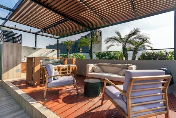 A rooftop terrace with a wooden deck, a white sofa, a wooden table with chairs, and a small round table.