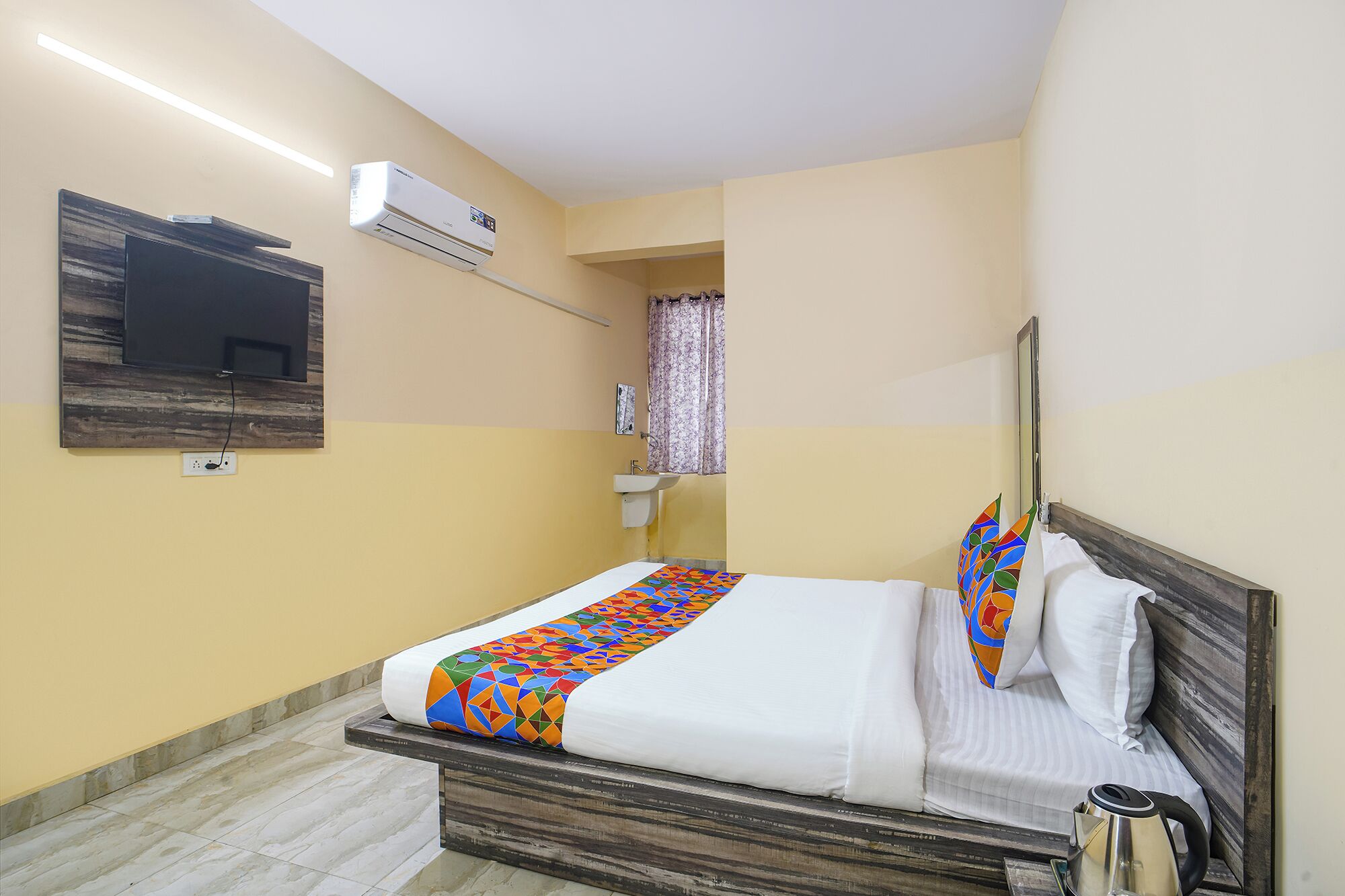 A hotel room with a bed, a TV mounted on the wall, an air conditioning unit, a sink, and a kettle on the bedside table.