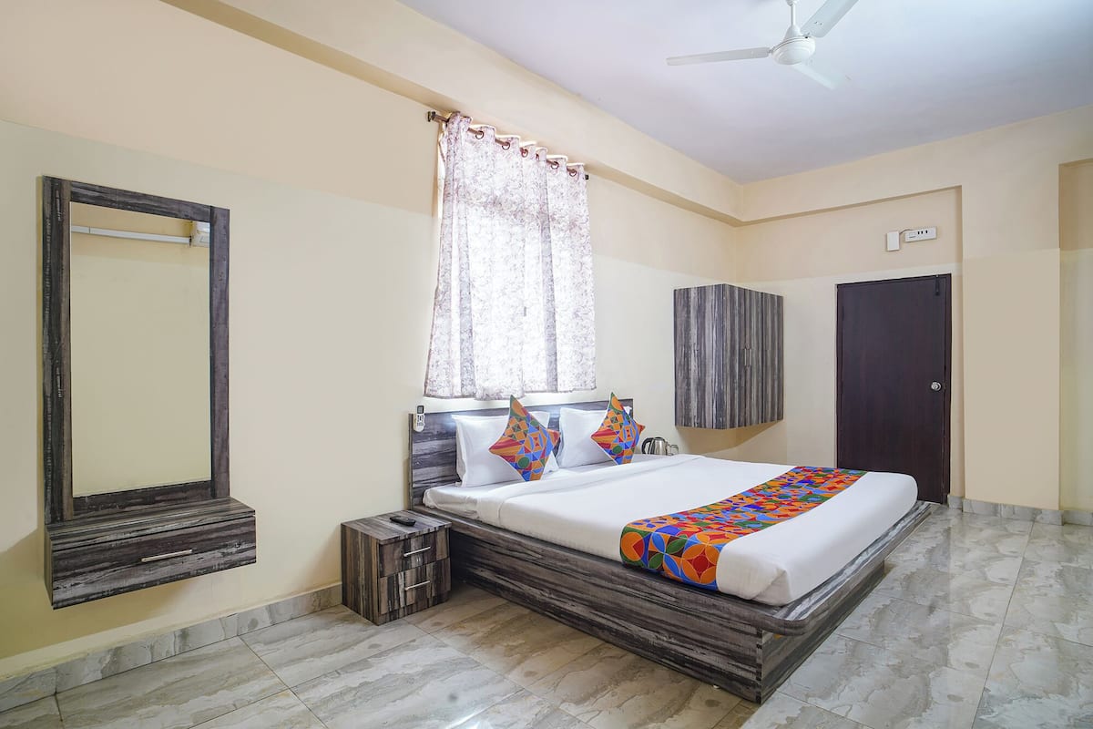 A bedroom with a bed, a nightstand, a mirror, a window with curtains, a ceiling fan, a wardrobe, and a door.