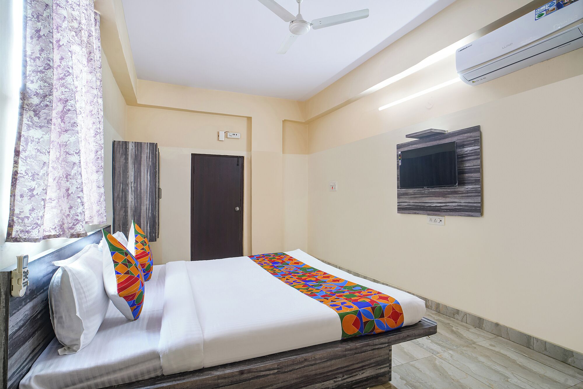 Camera d'albergo con un letto, una ventola a soffitto, un condizionatore d'aria, una porta e una TV a parete.