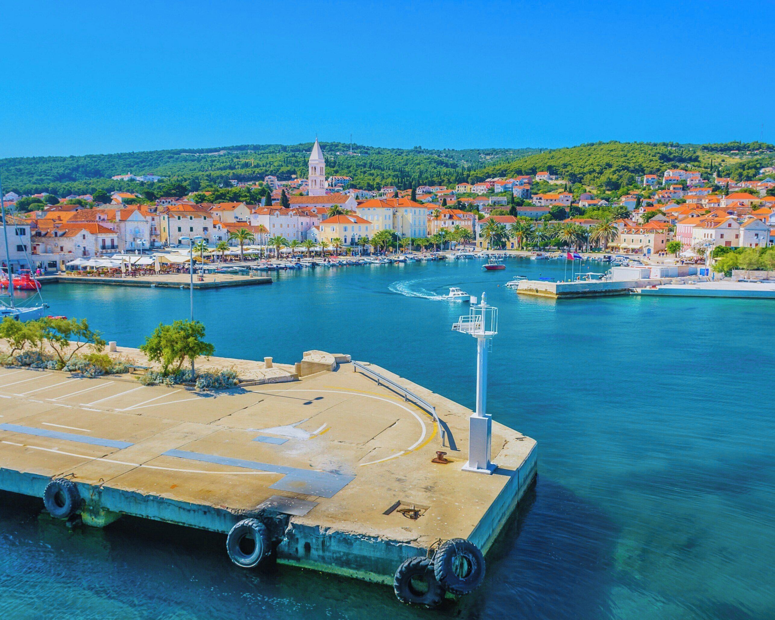 A waterfront marina with boats, a dock, and a town with red-roofed buildings in the background.
