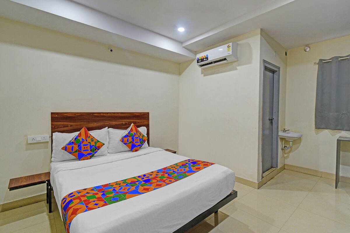 A hotel room with a bed, a bedside table, a wall-mounted air conditioner, a door, and a bathroom with a sink.