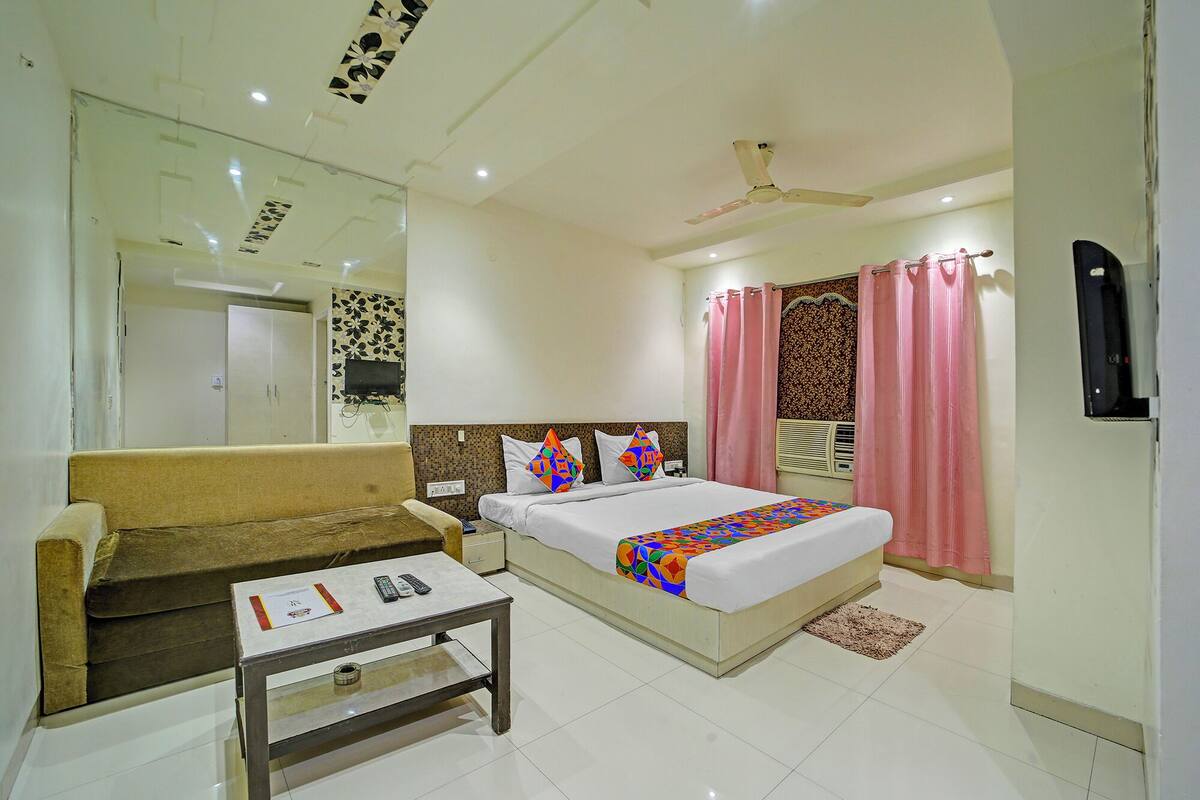 A hotel room with a bed, a sofa, a small table, a mirror, a ceiling fan, and pink curtains.