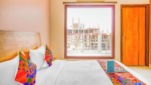 Une chambre d’hôtel avec une grande fenêtre, un lit avec du linge de maison blanc, des coussins colorés et une porte en bois.