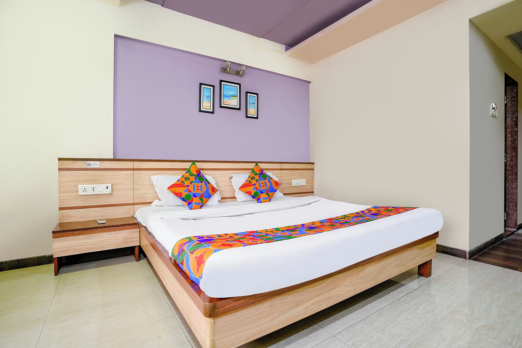 A hotel room with a wooden bed, white bedding, a purple accent wall, and a small bedside table.