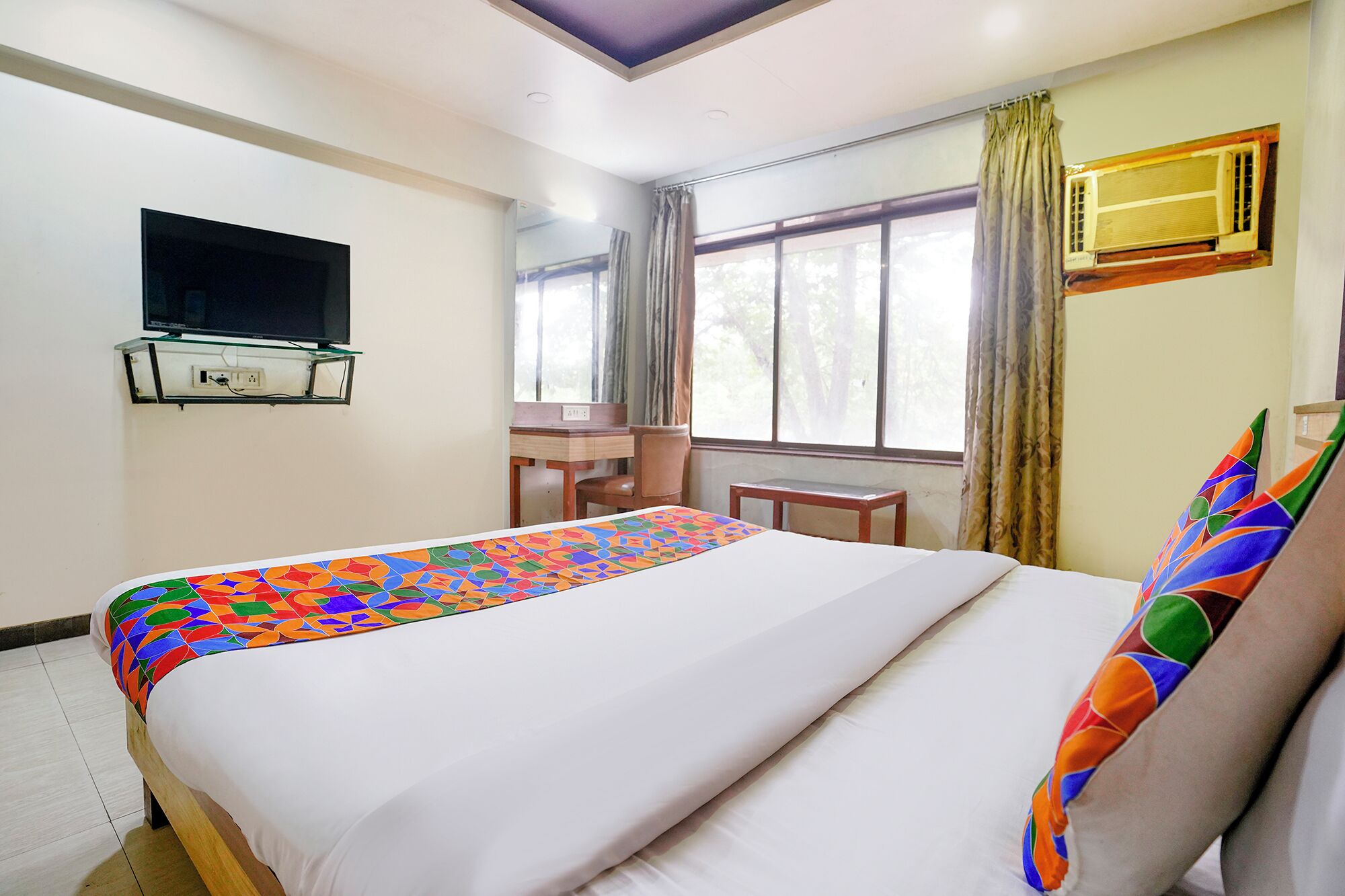 Habitación de hotel con cama, escritorio, silla, televisión y ventana con cortinas.