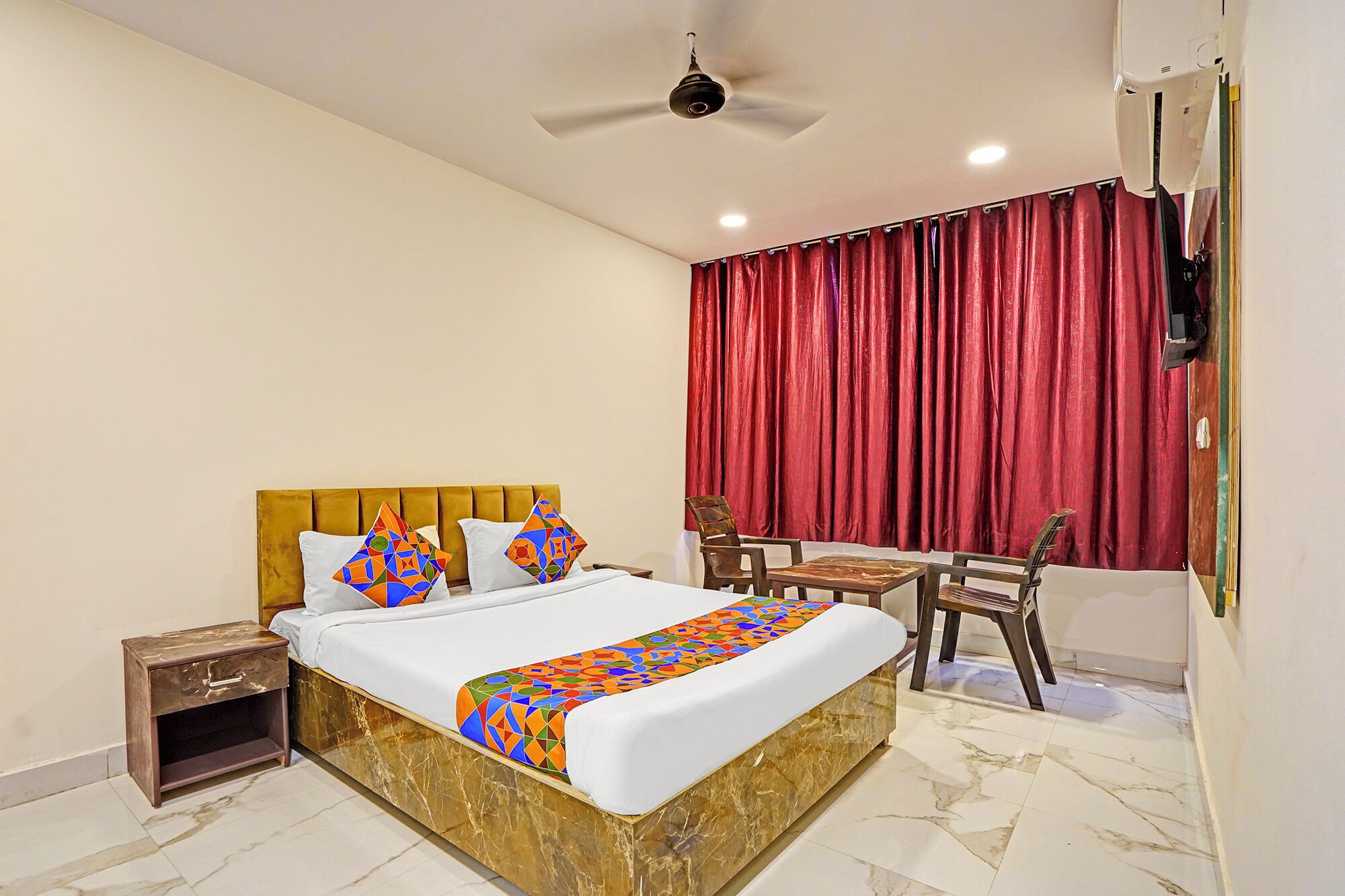A hotel room with a bed, a small table, a chair, a ceiling fan, and a window with red curtains.
