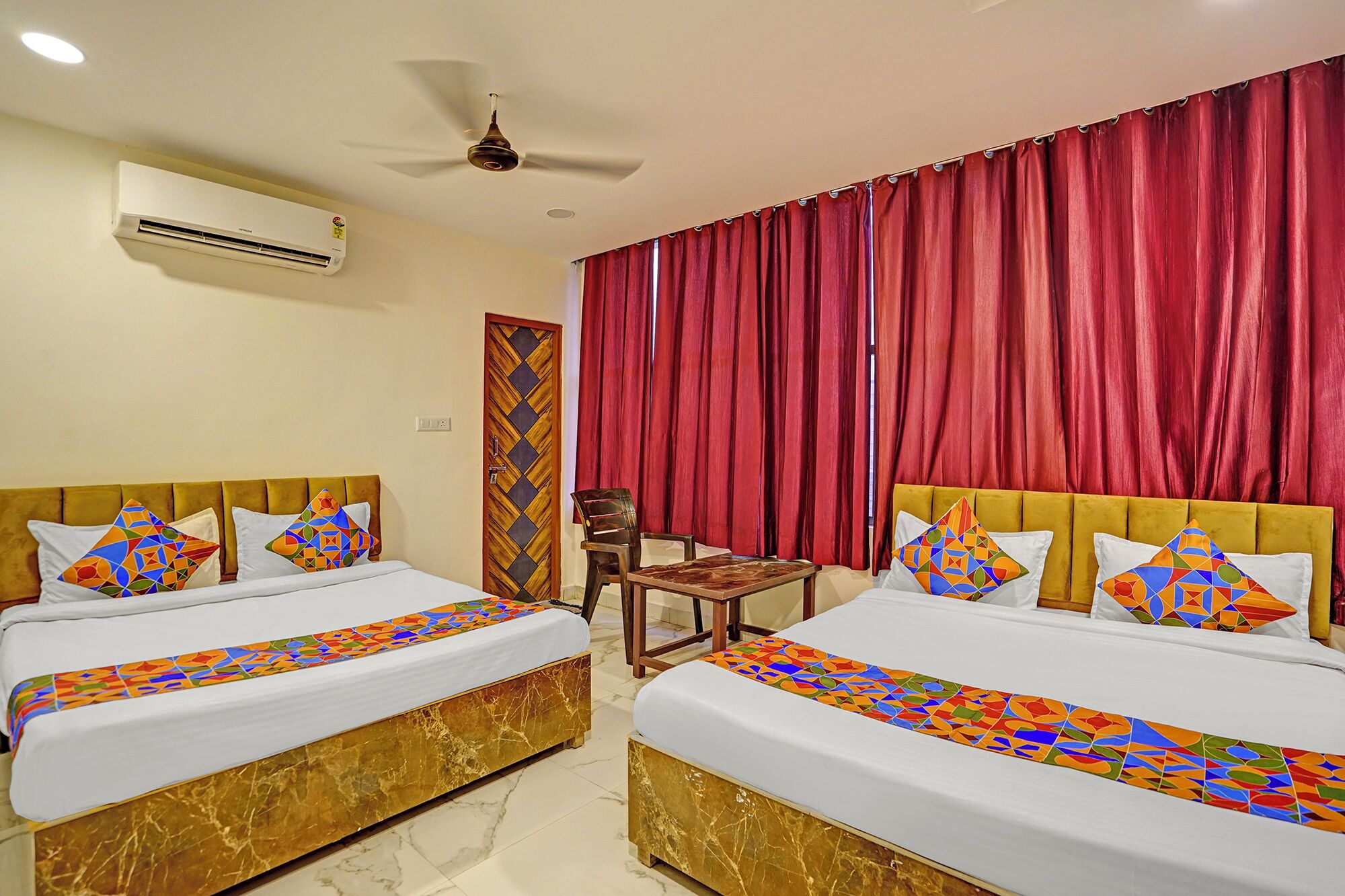 A hotel room with two beds, a ceiling fan, an air conditioning unit, a wooden door, and red curtains.