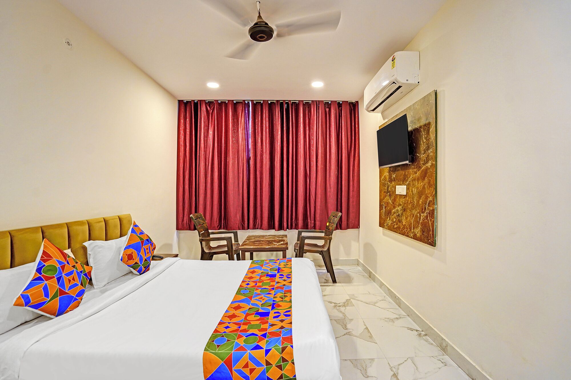 A hotel room with a bed, a ceiling fan, a TV mounted on the wall, and a window with red curtains.