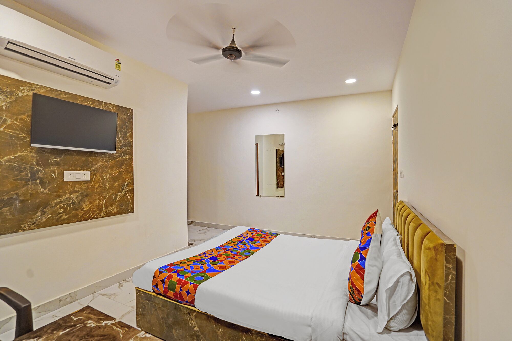 Una habitación de hotel moderna con suelo de mármol, una cama con colcha geométrica de colores, un televisor de pantalla plana en la pared y un ventilador de techo.