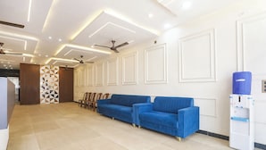 A modern waiting area with a blue sofa, ceiling fans, recessed lighting, and a water dispenser. - Fabhotel Shri Ram Retreat (Mathura)