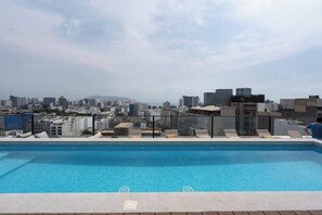 Piscina en la azotea con vista a la ciudad, sillas de descanso y barandilla de vidrio.