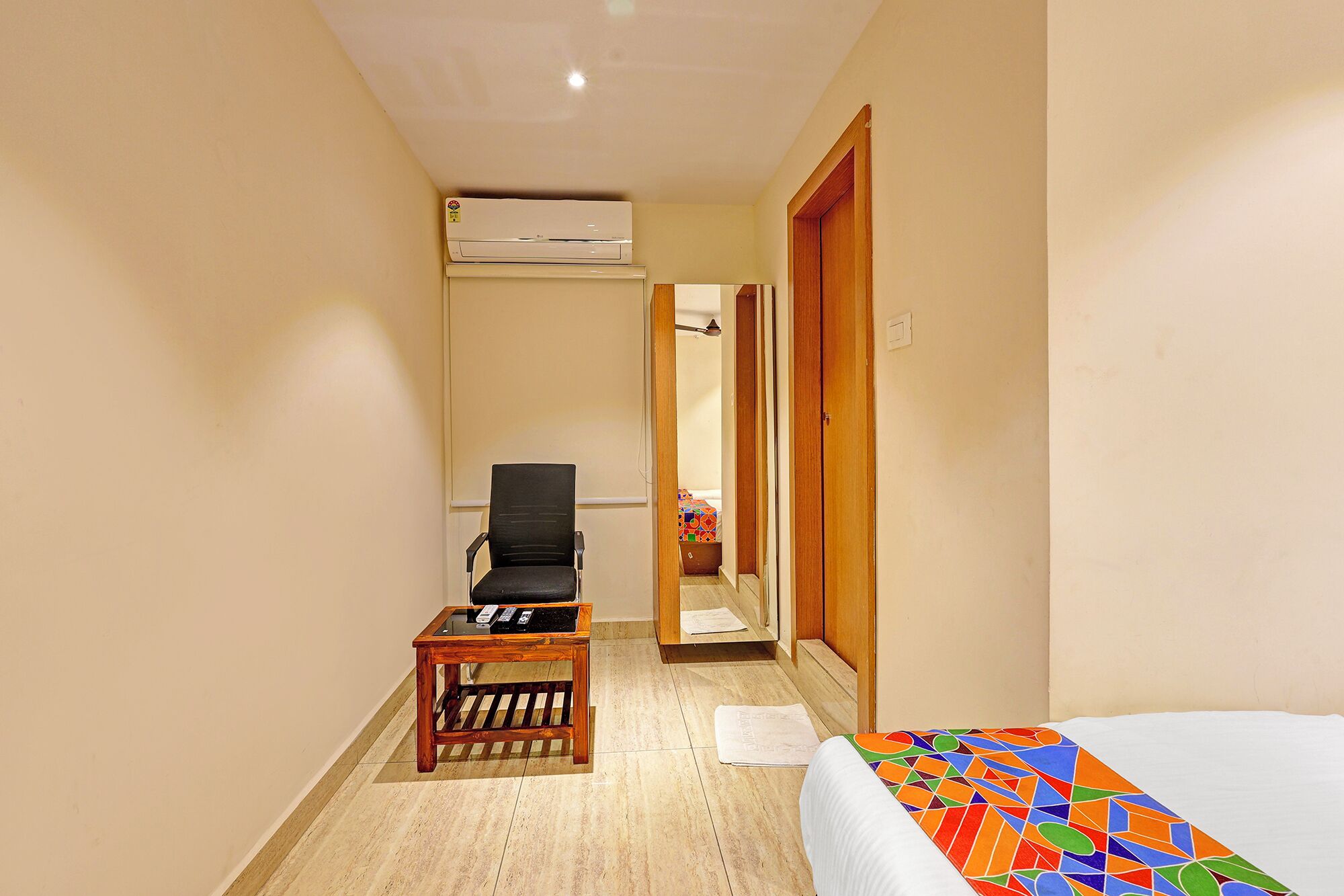 A hotel room with a bed, a chair, a small table, an air conditioning unit, and a door leading to another room.