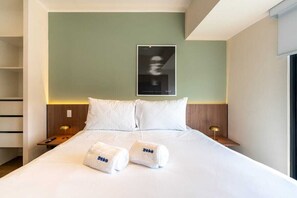 Een hotelkamer met een bed, twee kussens, twee nachtkastjes met lampen, een lijst aan de muur, en twee handdoeken met het merk 'geko' op het bed.