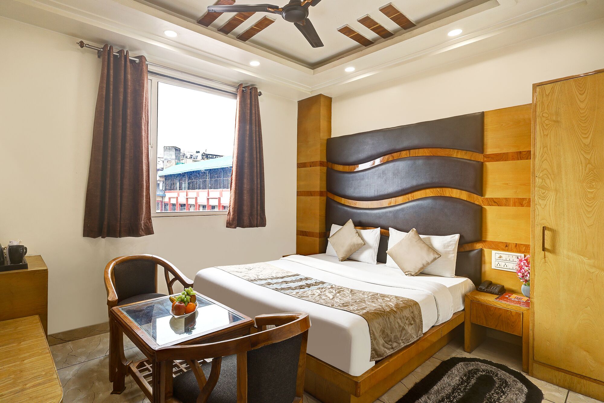 A hotel room with a bed, a desk, a chair, a window with curtains, a ceiling fan, and a view of buildings outside.