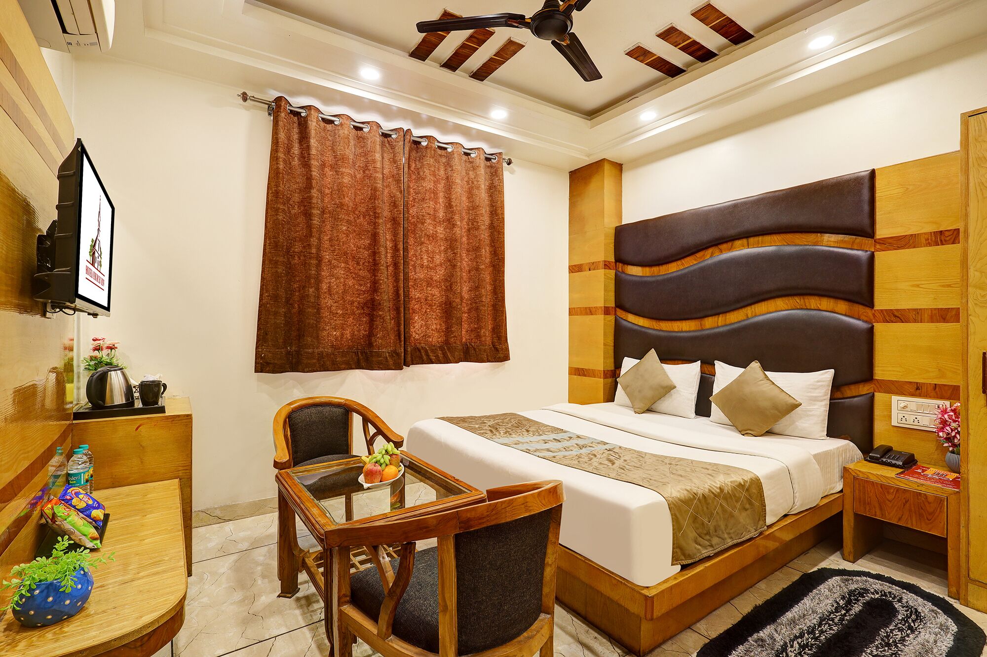 A hotel room with a bed, a wooden desk, a chair, a small table with a fruit bowl, a ceiling fan, and a wall-mounted TV.