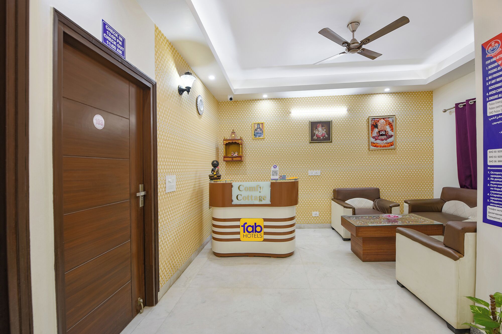 A reception area with a wooden reception desk labeled "Comfy Cottage" and "fab hotels". There are two sofas and a small table with a decorative item. A ceiling fan is visible above.
