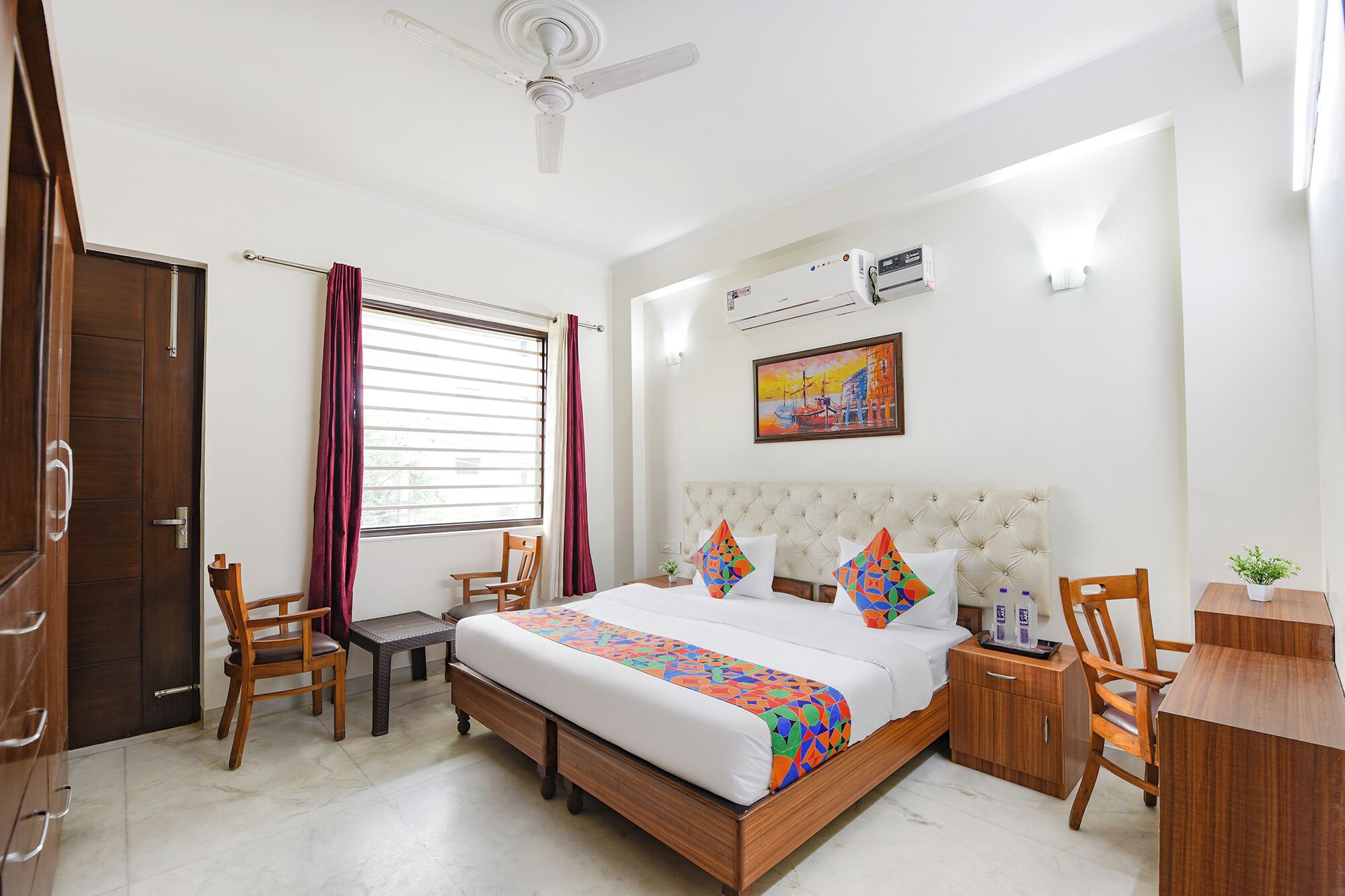 Habitación de hotel con cama, escritorio, silla, armario, ventana con cortinas, ventilador de techo, aire acondicionado y un cuadro en la pared.