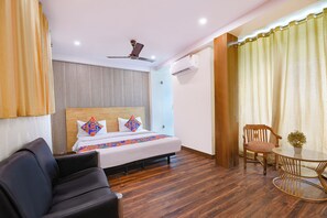 A hotel room with a bed, a ceiling fan, an air conditioning unit, a wooden chair, a small round table, and a potted plant.