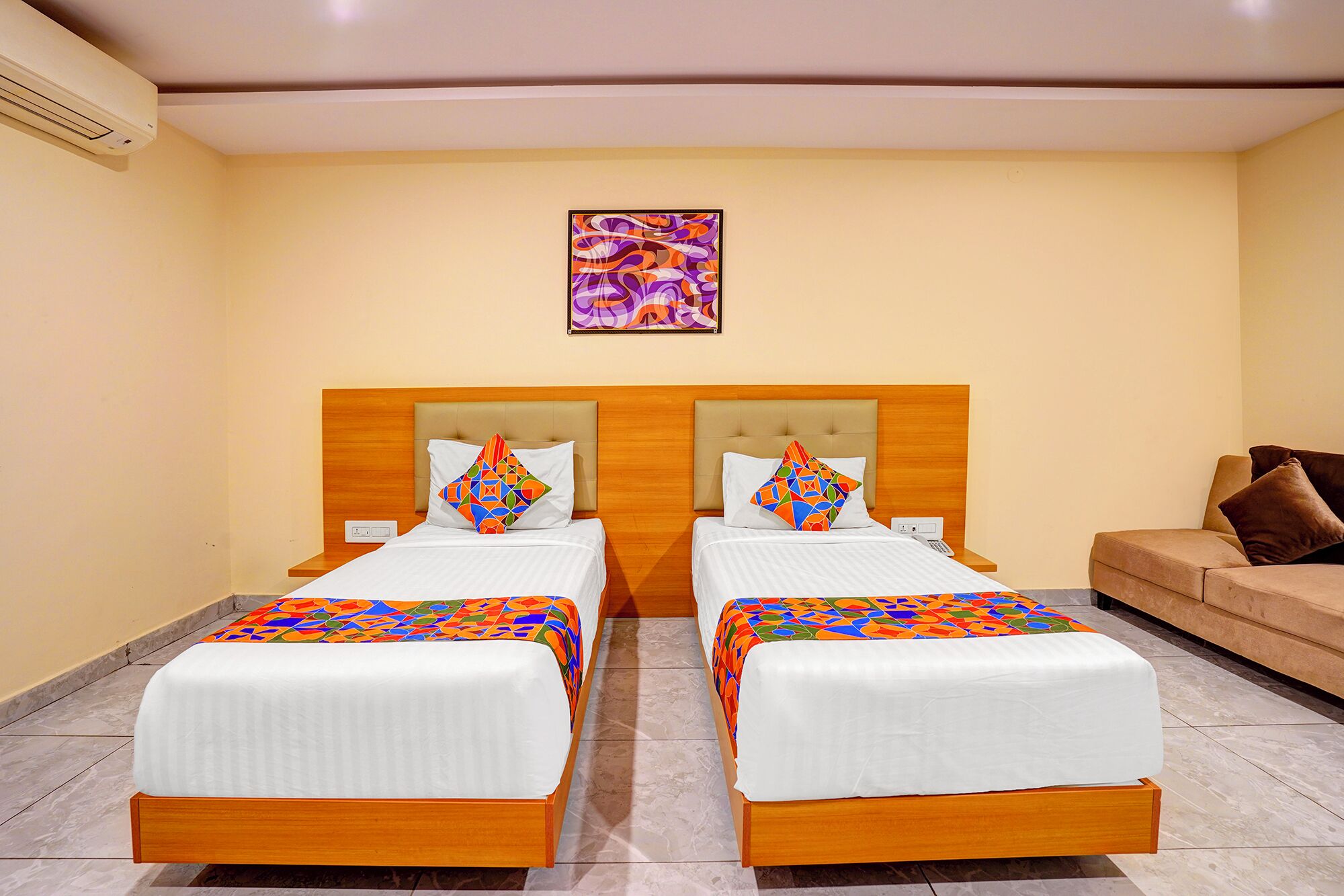 A hotel room with two beds, each with a colorful geometric-patterned bedspread and matching pillows. There is a single bed on the left and a sofa on the right.