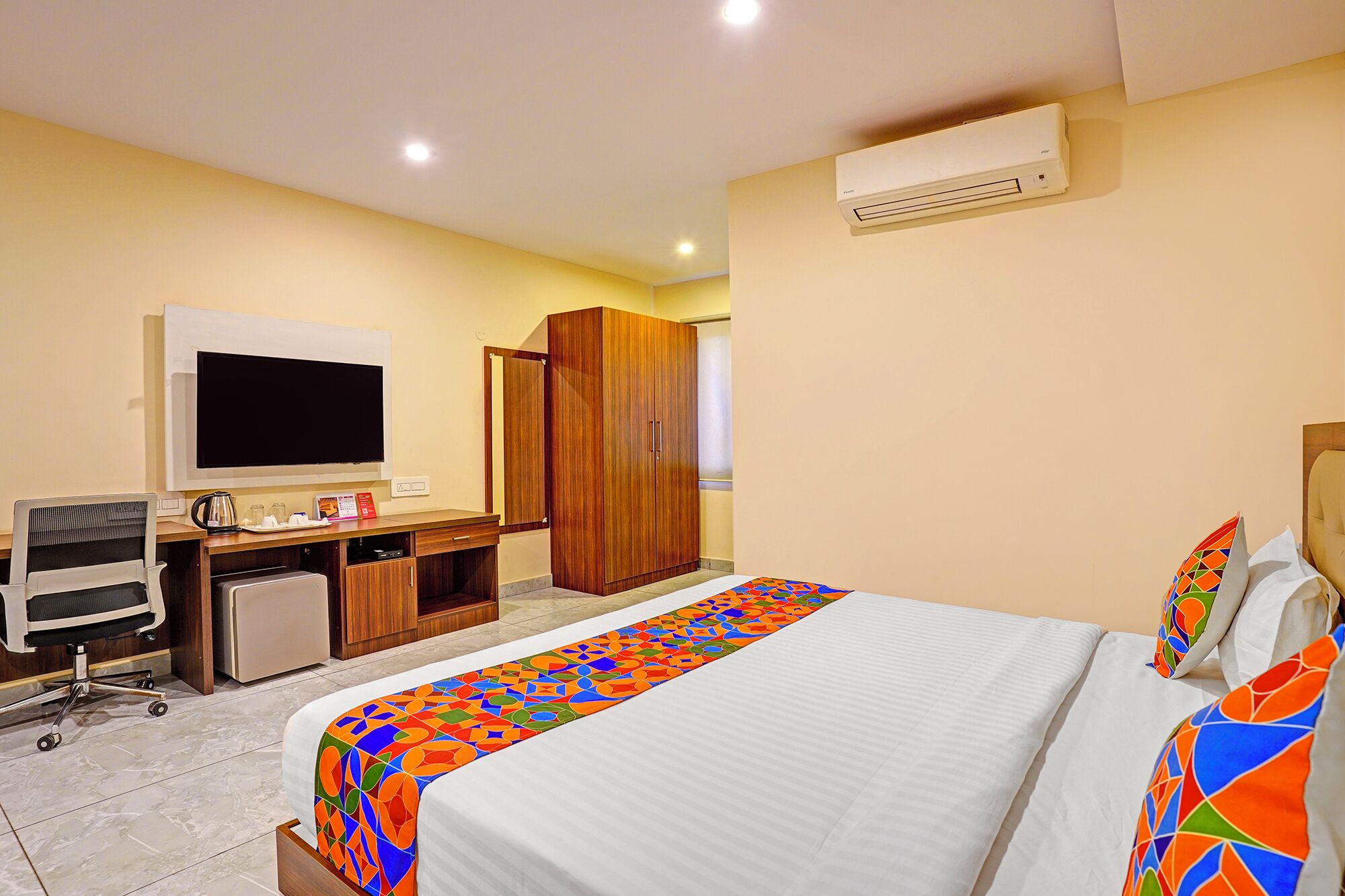A hotel room with a bed, a desk with a computer, a TV, and an air conditioning unit.