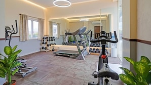 A well-lit gym with exercise equipment, including treadmills and dumbbells, and large windows with curtains. - FabHotel Millennials - Jigani (Anekal)