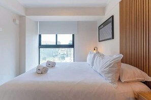 Hotellihuone, jossa on suuri sänky, ikkuna, josta avautuu näkymä kaupunkiin, puupaneloitu seinä ja kehystetty taideteos.
