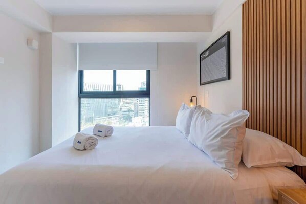 A hotel room with a large bed, a window with a city view, a wooden panel wall, and a framed artwork.