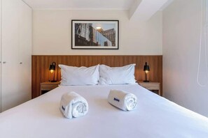 Hotellihuone, jossa on sänky, kaksi yöpöytälamppua, seinällä oleva kehystetty kuva ja kaksi rullalle taitettua pyyhettä sängyllä.