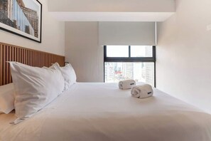 Hotellihuone, jossa on sänky, tyynyjä, ikkuna, josta on näkymä rakennuksiin, ja seinällä oleva kehystetty kuva.