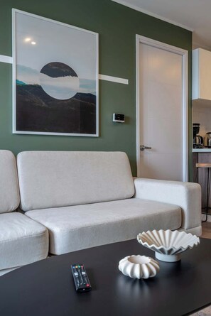 Un salon comprenant un canapé, une table basse avec une télécommande et des objets décoratifs, un tableau encadré au mur et une porte.