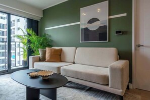 Un salon avec un canapé, une table basse, une grande fenêtre et un mur vert orné d’un tableau.