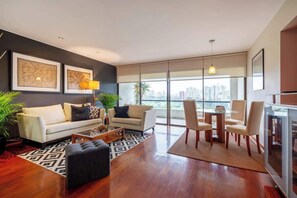 A modern living room with a sofa, coffee table, and chairs, large windows with a city view, and artwork on the walls. - Geko | Inviting 2BR in San Isidro w/ Park Views (San Isidro)