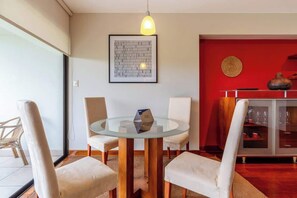 A dining area with a glass table, four chairs, a red accent wall, a framed artwork, and a cabinet with glass doors. - Geko | Inviting 2BR in San Isidro w/ Park Views (San Isidro)