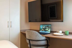 Een moderne hotelkamer met een flatscreen televisie aan de muur, een laptop op een bureau, een stoel, een bed en een klein tafeltje met een kop en schotel.