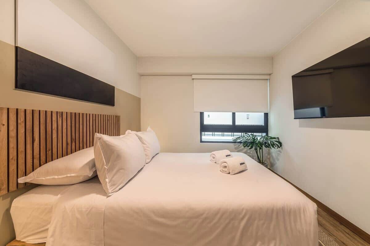 A hotel room with a bed, pillows, a TV, a window, and a plant.