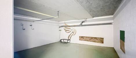 A parking garage with concrete floors and walls, fluorescent lighting, and visible pipes.