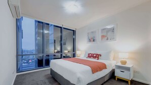 A modern bedroom with a large bed, a nightstand, a window with a city view, and a ceiling light.
