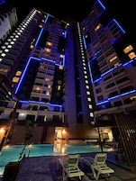 Un edificio de gran altura con líneas azules iluminadas y un área de piscina con sillones de descanso.
