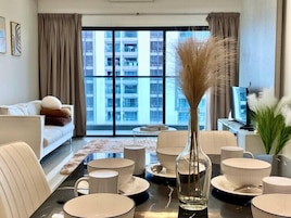 Una sala de estar moderna con un ventanal grande, un sofá, una mesa de comedor con platos y tazas blancos, y un florero con plantas secas.