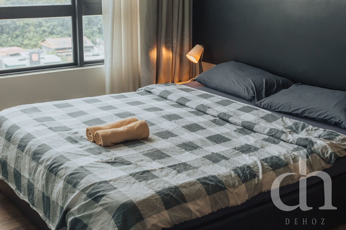 A neatly made bed with a checkered bedspread and a rolled towel, a bedside lamp, and a view of the outside through a window.
