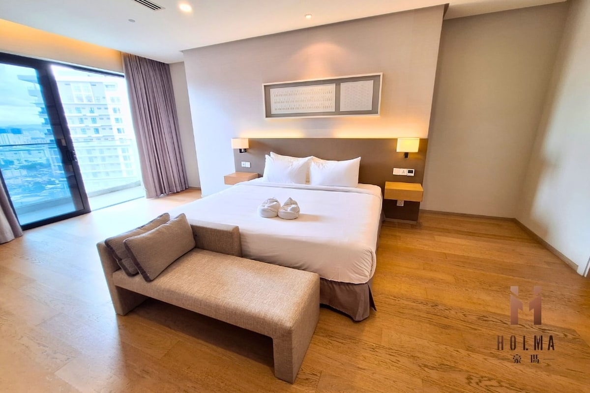 A hotel room with a large bed, two bedside tables, a bench, a large window with a view of buildings, and a framed artwork on the wall.