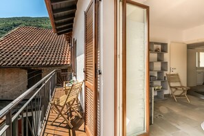 A balcony with a tiled roof, a wooden table and chairs, and a sliding glass door leading to a room with shelves and a chair.