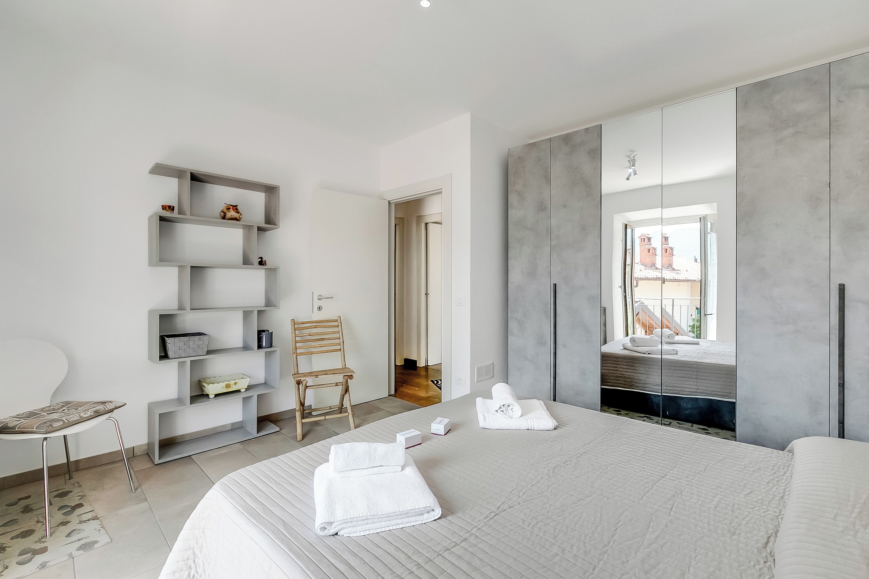 A modern hotel room with a large bed, a built-in wardrobe, a seating area with a chair, and a view of the outside.