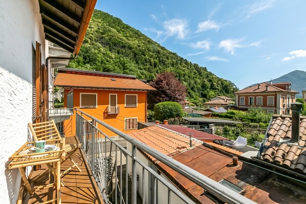 A balcony with a view of a mountain, a terrace with a table and chairs, and a building with a red roof.